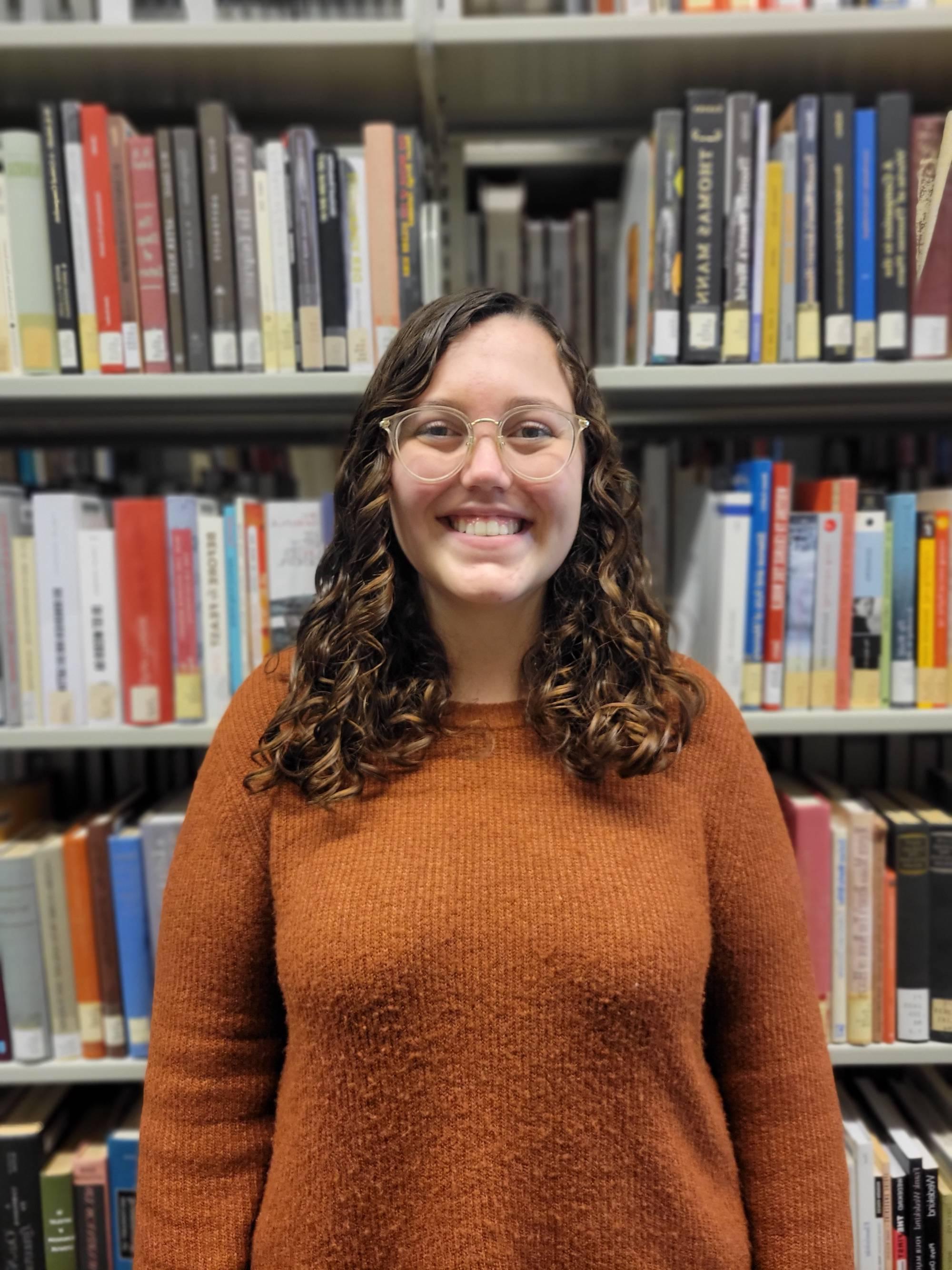 Smiling woman with curly brown hair wearing a yellow sweater with books in the background